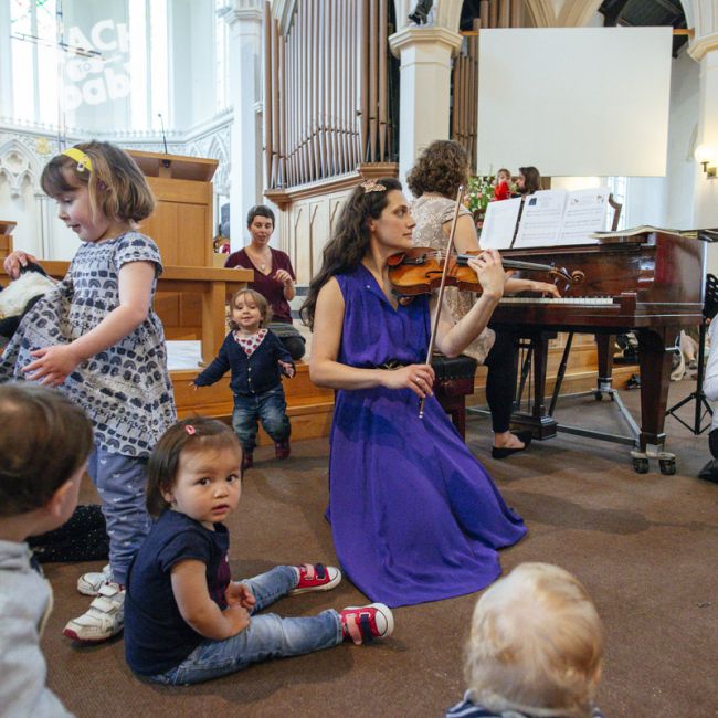 A violinist wearing a blue dress playing the violin in a church, kneeling on the floor. There are toddlers and young children sitting and standing to her left